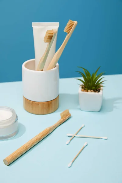 Soporte con pasta de dientes en tubo y cepillos de dientes de bambú, crema cosmética y planta en maceta sobre mesa y fondo azul - foto de stock