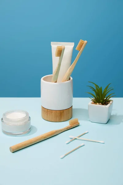 Titular com pasta de dentes em tubo e escovas de bambu, creme cosmético e planta em vaso na mesa e fundo azul — Fotografia de Stock