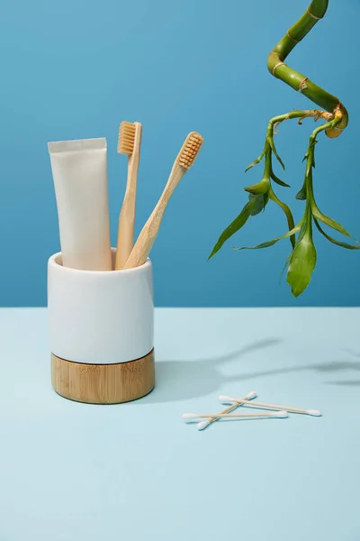 Тримач з бамбуковими зубними щітками, зубна паста в трубці, вушні палички і бамбуковий стебло на столі і синій фон — стокове фото