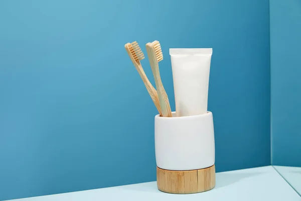 Holder with bamboo toothbrushes, toothpaste in tube and mirror on table and blue background — Stock Photo