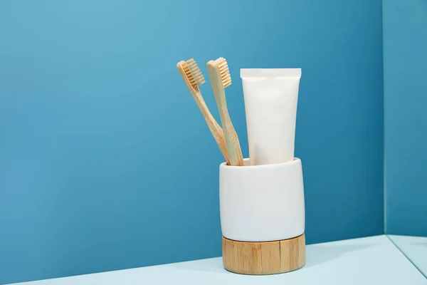 Soporte con cepillos de dientes de bambú, pasta de dientes en tubo y espejo sobre mesa y fondo azul - foto de stock