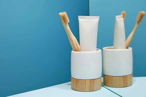 Support avec brosses à dents en bambou, dentifrice en tube près du miroir sur table et fond bleu — Photo de stock