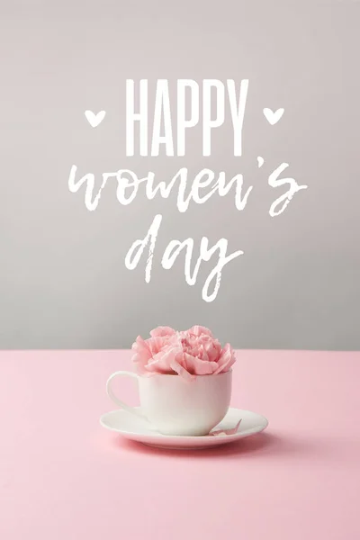 Розовые гвоздичные цветы в белой чашке на блюдце на сером фоне с счастливым женским днем надписи — стоковое фото
