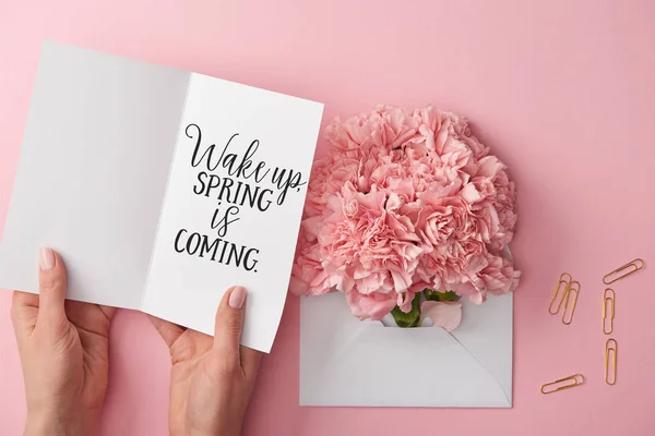 Cropped view of woman holding greeting card with wake up, spring is coming lettering near carnation flowers in envelope on pink background — Stock Photo