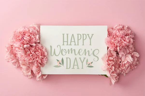 Vista superior do cartão de saudação com letras do dia feliz das mulheres e cravos rosa no fundo rosa — Fotografia de Stock