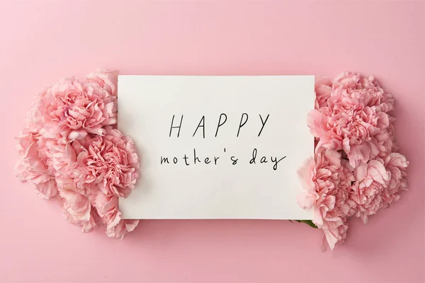 Vista superior de la tarjeta de felicitación con letras feliz día de las madres y claveles de color rosa sobre fondo rosa - foto de stock