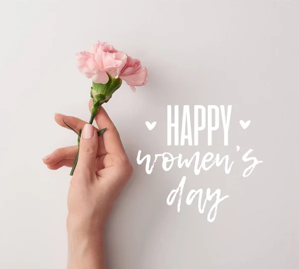 Partial view of woman holding pink carnation on grey background with happy womens day lettering — Stock Photo