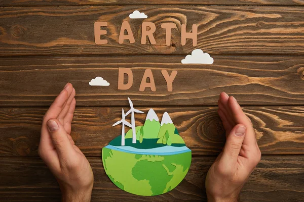 Teilansicht einer Frau, die einen aus Papier geschnittenen Planeten mit erneuerbaren Energiequellen und Buchstaben auf Holzgrund hält, Earth Day Konzept — Stockfoto