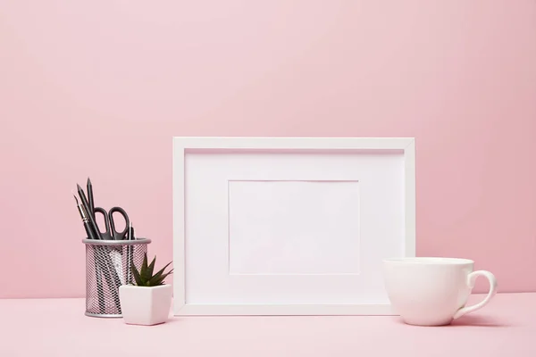 Blank frame near green plant, metallic holder with stationery and cup — Stock Photo