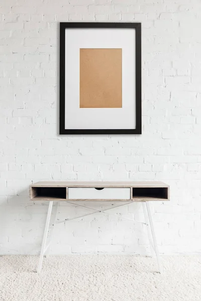 Modern table standing on carpet near black frame hanging on white brick wall — Stock Photo