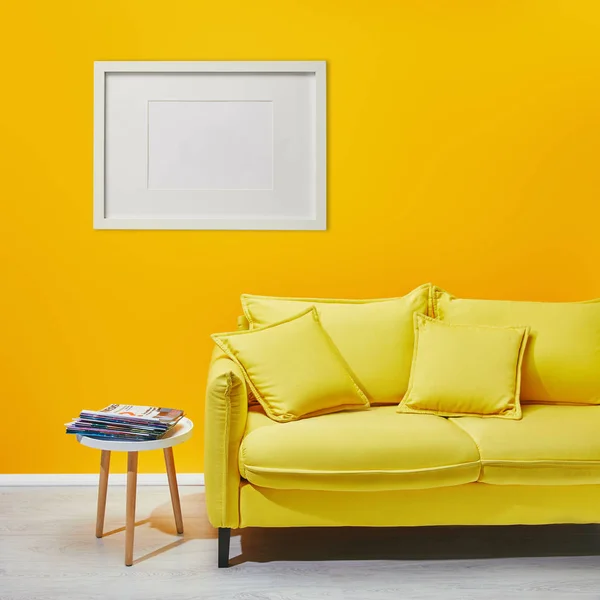 Mesa de centro de pie cerca de sofá amarillo moderno cerca de marco blanco colgando en la pared - foto de stock