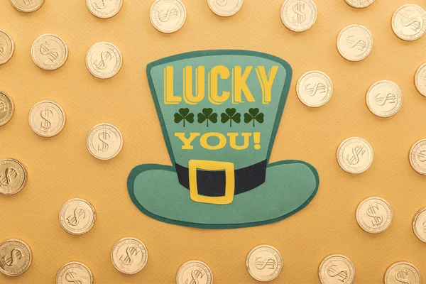 Top view of golden coins with dollar signs near green paper hat with lucky you lettering on orange background — Stock Photo