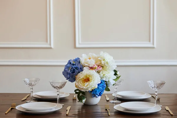 Piatti bianchi, coltelli e forchette dorate, bicchieri di cristallo e fiori in vaso sul tavolo di legno nel ristorante — Foto stock