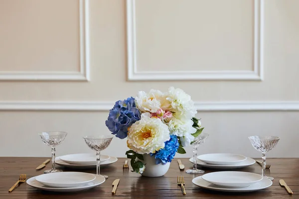 White plates, golden knifes and forks, crystal glasses and flowers in vase on wooden table in restaurant — Stock Photo