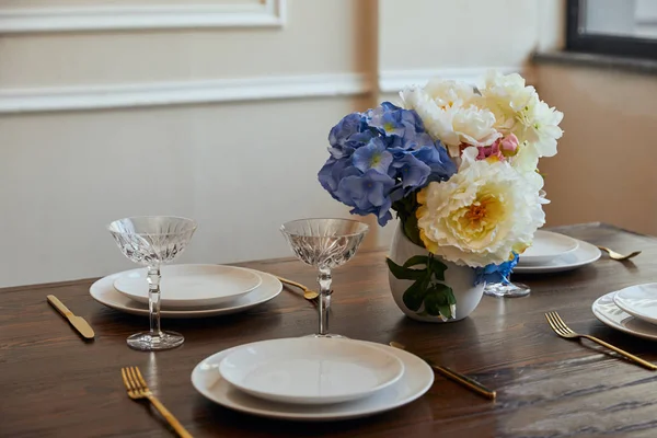 Assiettes blanches, couteaux et fourchettes, verres en cristal et fleurs en vase sur table en bois à la maison — Photo de stock