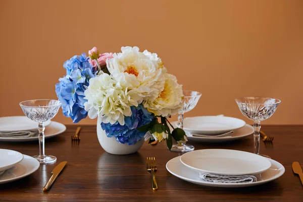 Piatti, coltelli e forchette dorati, bicchieri di cristallo e bouquet in vaso bianco su tavolo di legno isolato su beige — Foto stock