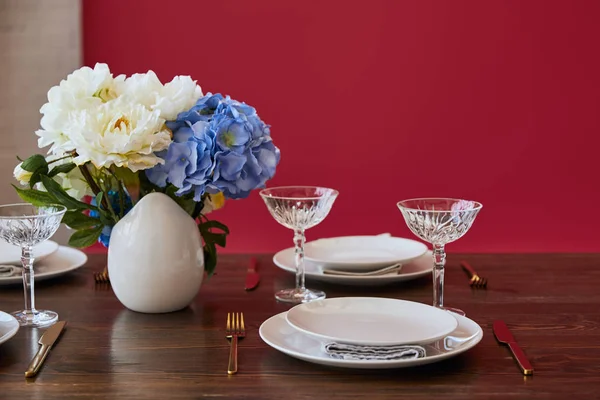 Assiettes, couteaux et fourchettes dorés, verres en cristal et bouquet en vase blanc sur table en bois près du mur rouge à la maison — Photo de stock