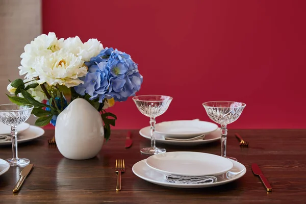 Plates, golden knifes and forks, crystal glasses and bouquet in white vase on wooden table near red wall at home — Stock Photo