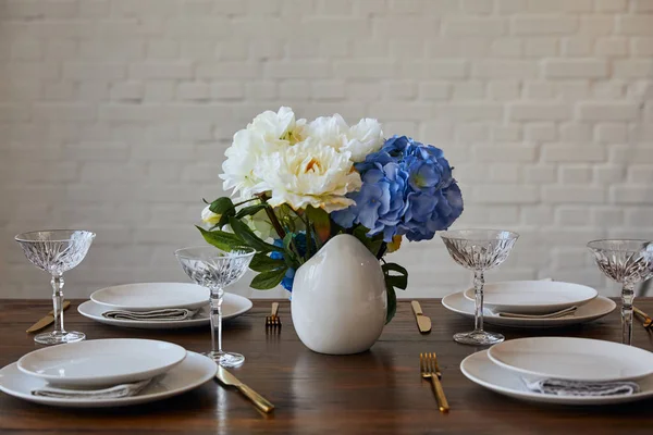 Assiettes, couteaux et fourchettes dorés, verres en cristal et bouquet en vase blanc sur table en bois près du mur de briques à la maison — Photo de stock