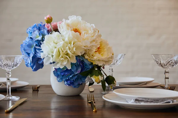 Assiettes, couteaux et fourchettes, verres en cristal et bouquet en vase blanc sur table en bois près du mur de briques à la maison — Photo de stock