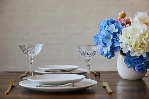 Piatti bianchi, coltelli e forchette, bicchieri di cristallo e bouquet in vaso sul tavolo di legno vicino al muro di mattoni a casa — Foto stock