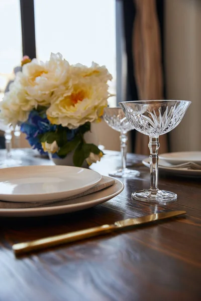 Weiße Teller, Kristallgläser und Strauß in weißer Vase auf Holztisch zu Hause — Stockfoto