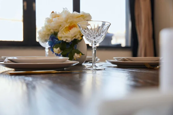 Pratos, copos de cristal e flores em vaso branco na mesa de madeira em casa — Fotografia de Stock