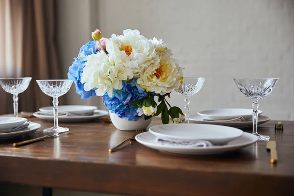 Assiettes blanches, couverts, verres en cristal et bouquet en vase sur table en bois à la maison — Photo de stock