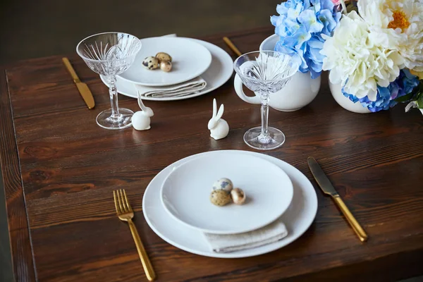 Verre cristal, fleurs dans un vase, couverts dorés et lapins décoratifs sur table en bois à la maison — Photo de stock