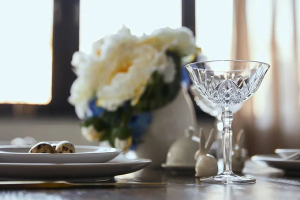 Messa a fuoco selettiva di vetro cristallo, bouquet in vaso e conigli decorativi su tavolo in legno a casa — Foto stock