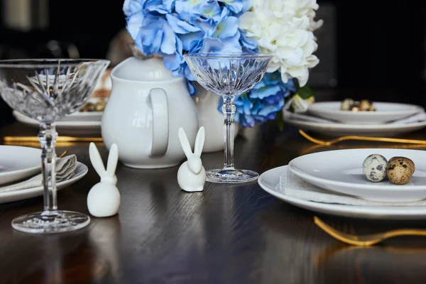 Foyer sélectif de lapins décoratifs près des assiettes, verre de cristal et fleurs sur table en bois à la maison — Photo de stock