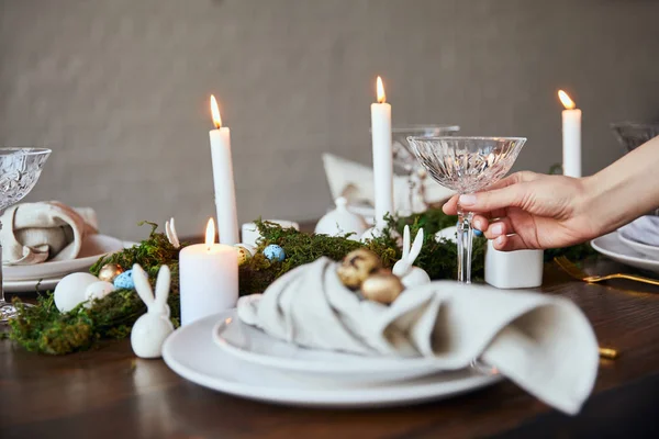 Cropped view of woman putting crystal glass near plates, burning candles and moss on wooden table at home — Stock Photo