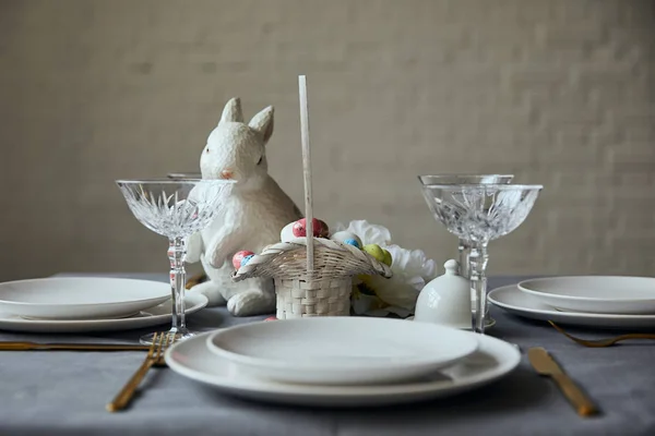 Белые тарелки, столовые приборы, хрустальные бокалы, декоративный кролик и корзина с покрашенными яйцами на столе дома — стоковое фото