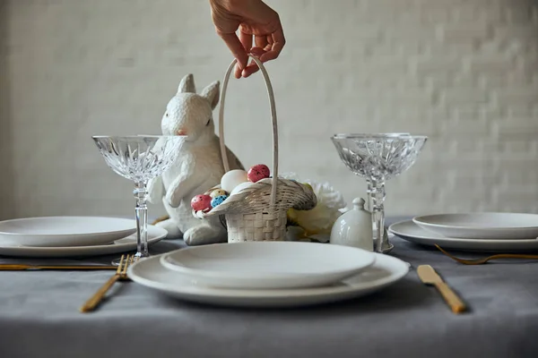 Vista ritagliata di donna mettendo cesto con uova dipinte vicino piatti bianchi, bicchieri di cristallo e coniglietto decorativo sul tavolo a casa — Foto stock