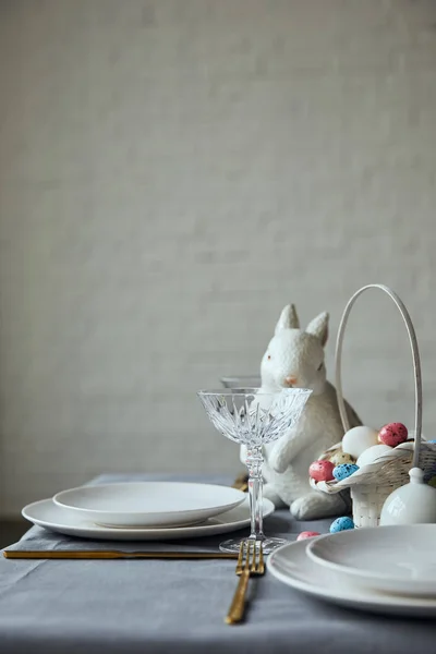 Placas brancas, vidro de cristal, coelho decorativo e cesta com ovos pintados na mesa em casa — Fotografia de Stock