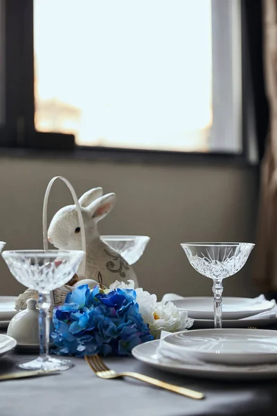 Platos, vidrio de cristal, conejito decorativo y flores en la mesa en casa - foto de stock