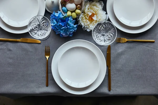 Vue de dessus des assiettes blanches, couverts, verres en cristal, panier avec des œufs peints et couverts sur la table à la maison — Photo de stock