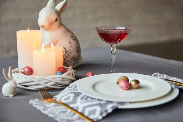 Foco seletivo de placas com guardanapo, ovos e velas em chamas na cesta, vinho em vidro cristal e coelho decorativo na mesa em casa — Fotografia de Stock