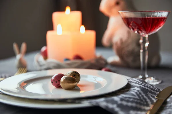 Messa a fuoco selettiva di piatti con uova, vino in cristallo, candele accese e coniglietti decorativi a tavola a casa — Foto stock