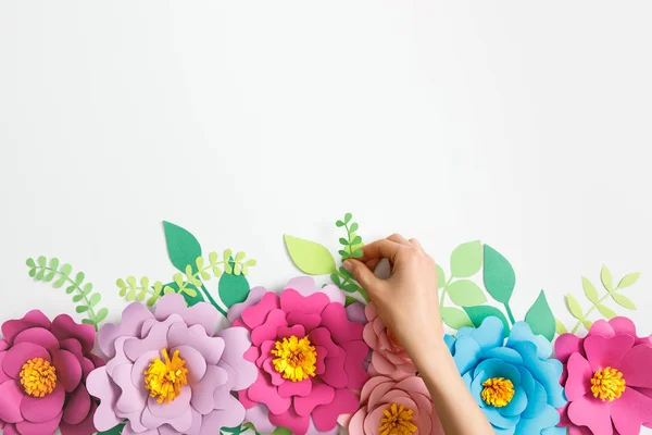 Vista recortada de la mujer poniendo planta verde cerca de flores de papel multicolor sobre fondo gris - foto de stock