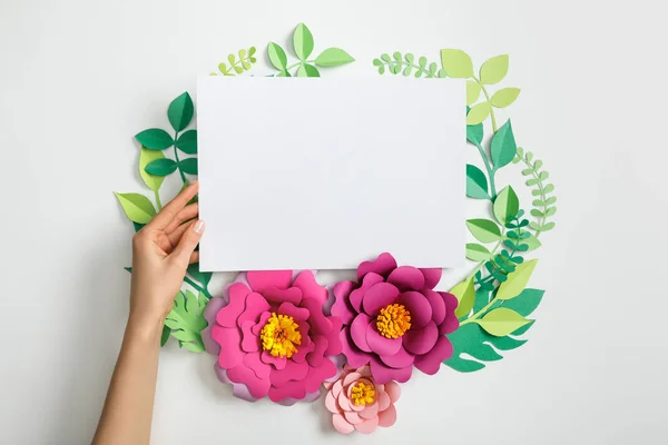 Vista recortada de la mujer poniendo blanco tarjeta en blanco cerca de flores de papel rosa y hojas sobre fondo gris - foto de stock