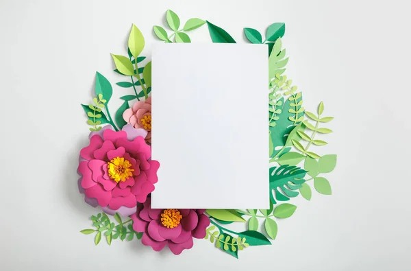 Top view of white blank card on paper flowers and green leaves on grey background — Stock Photo