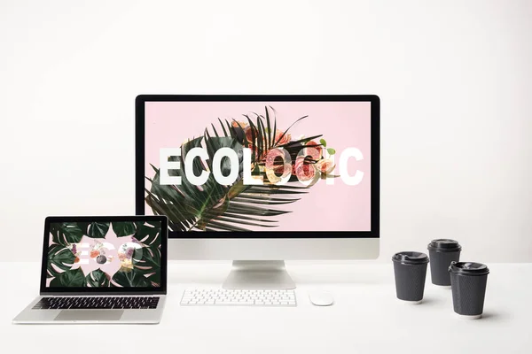Computer with ecologic lettering on monitor and laptop with eco lettering on screen on desk with paper cups on white background — Stock Photo