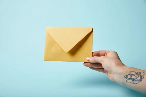 Partial view of woman with tattoo holding yellow envelope on blue background — Stock Photo