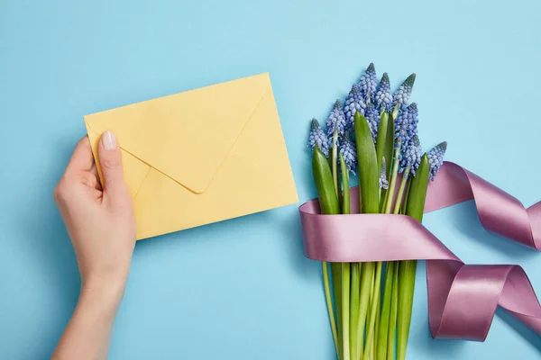 Vue partielle de la main femelle avec enveloppe jaune, et jacinthes bleues avec ruban de satin violet sur bleu — Photo de stock