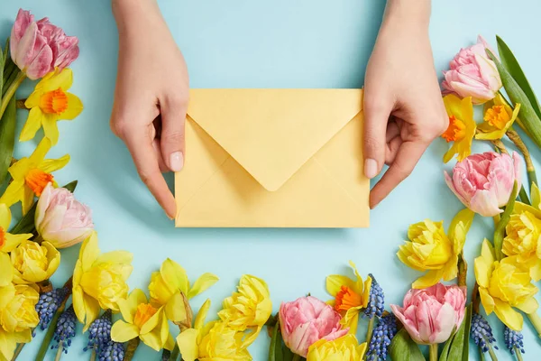 Vue partielle des mains féminines avec enveloppe jaune, tulipes roses, jonquilles jaunes et jacinthes bleues sur bleu — Photo de stock