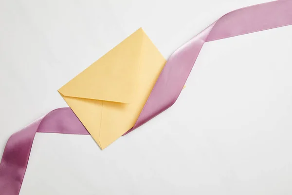 Вид зверху на жовтий конверт і фіолетову атласну стрічку на білому — стокове фото