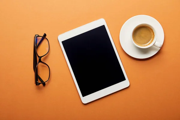 Вид сверху на цифровой планшет с пустым экраном рядом с чашкой кофе и стаканами на оранжевом — стоковое фото