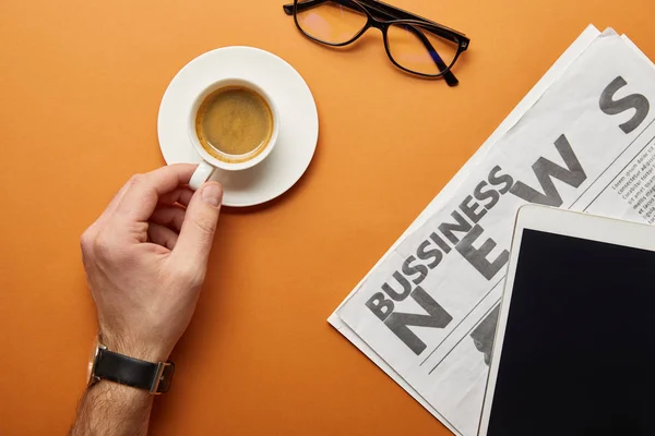 Vista recortada del hombre sosteniendo taza de café cerca de la tableta digital con pantalla en blanco, gafas y periódico de negocios en naranja - foto de stock