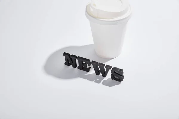 Black news lettering near paper cup with shadows on white — Stock Photo